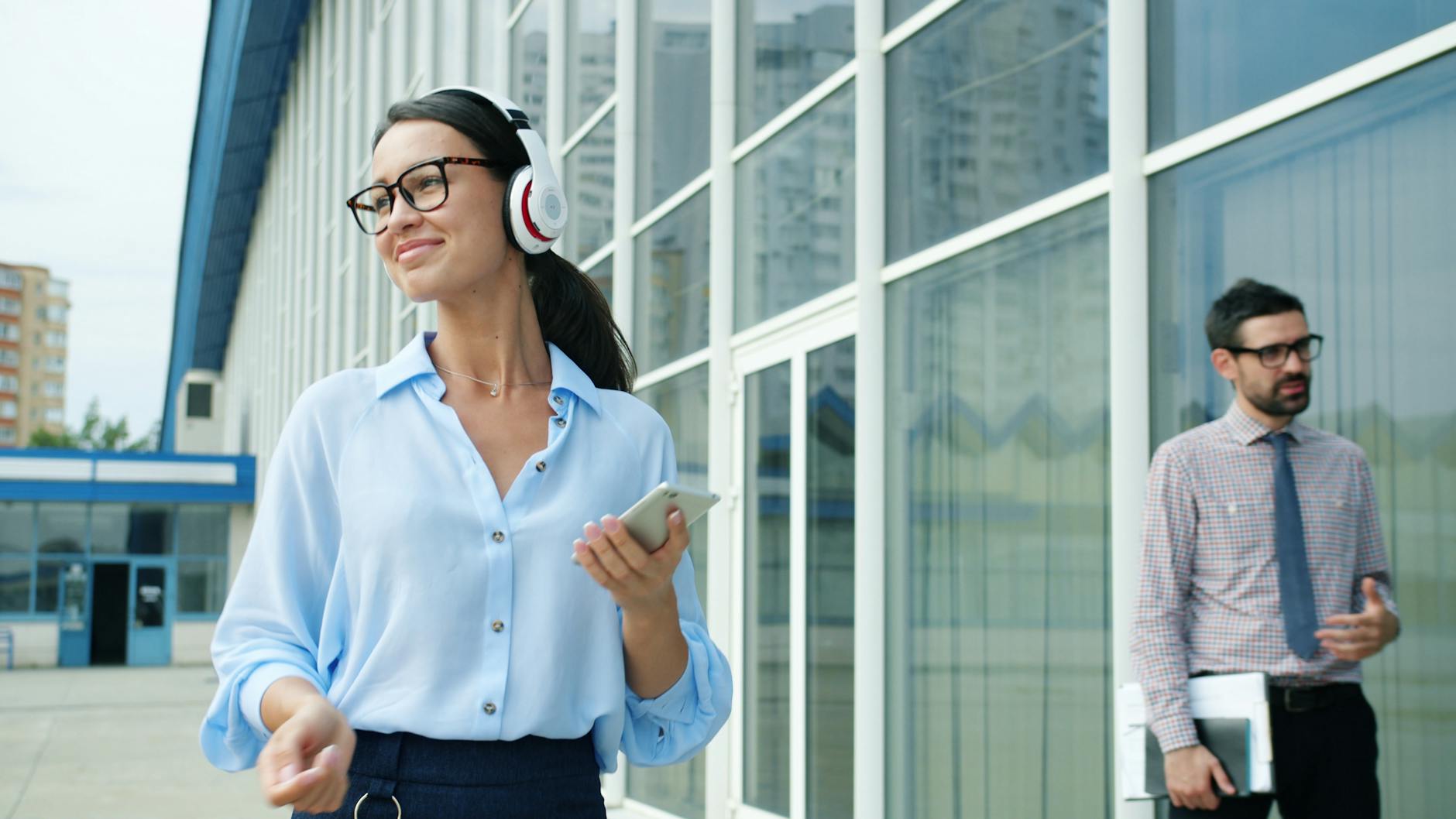 Uśmiechnięta pracownica biurowa słucha muzyki w słuchawkach przed biurowcem