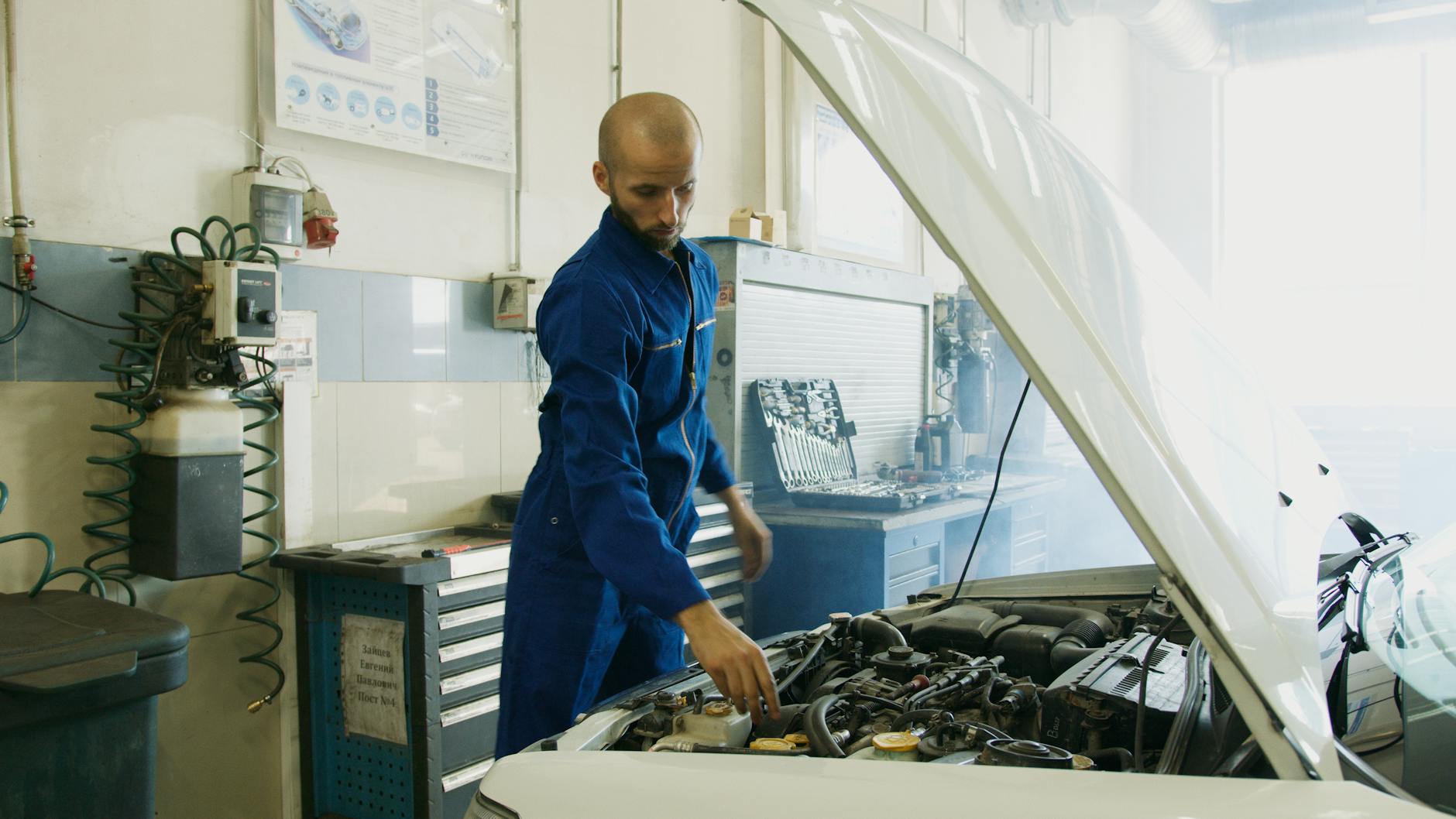 Mechanik w niebieskim kombinezonie sprawdza silnik auta w warsztacie Mechanik w niebieskim kombinezonie sprawdza silnik auta w warsztacie